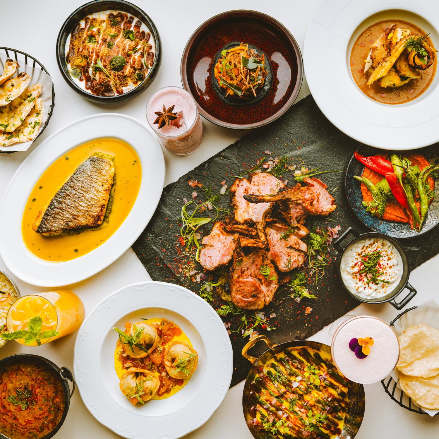 Table garnie d'un festin de plats gastronomiques variés, incluant du poisson en sauce, des grillades de viande, des légumes colorés, des sauces crémeuses et des boissons exotiques, présentés sur une nappe blanche avec une touche raffinée.
