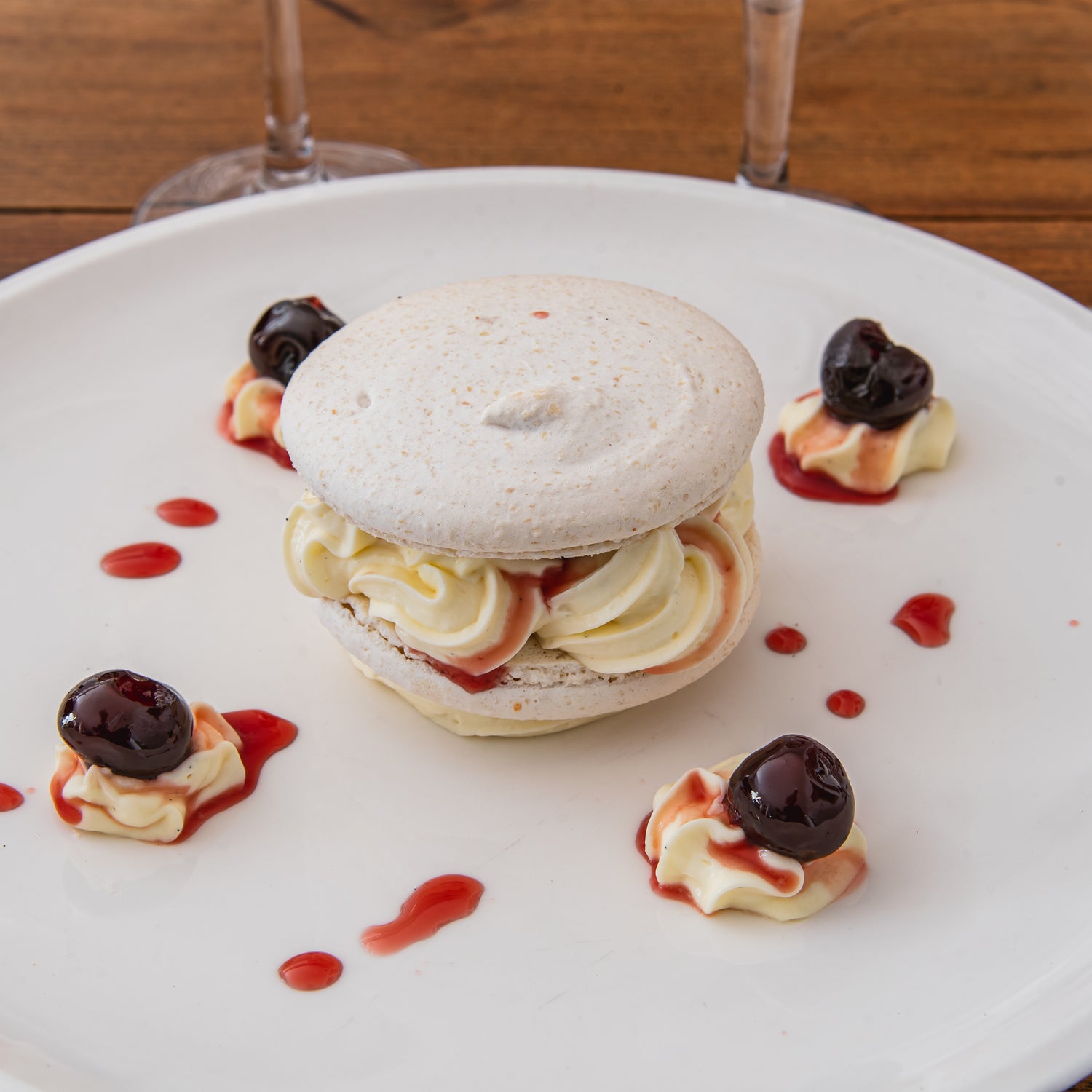 Dessert gourmand composé d’un macaron garni de crème vanillée et de coulis de fruits rouges, décoré de cerises confites sur une assiette blanche.