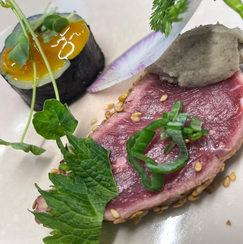 Assiette raffinée avec une pièce de thon mi-cuit en croûte de sésame, accompagnée d'une préparation noire garnie de gelée dorée et de fines herbes fraîches.