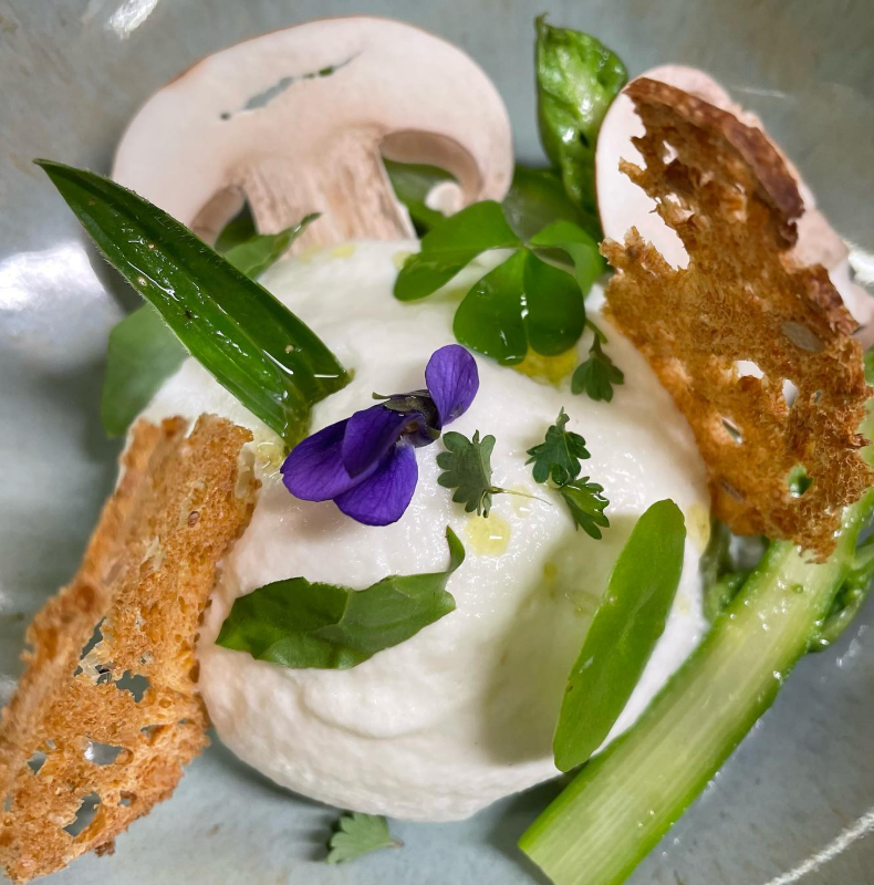 Plat gastronomique composé d'une mousse blanche accompagnée d'asperges vertes, de champignons, de croûtons dorés et décorée d'une fleur violette.