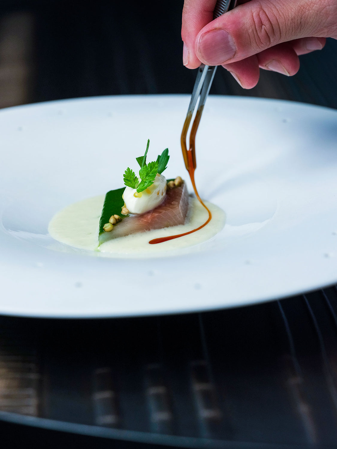 Gros plan sur un plat gastronomique : filet d'omble accompagné d'une sauce délicatement dressée, avec une main de chef ajoutant la touche finale à l'aide d'une pince.