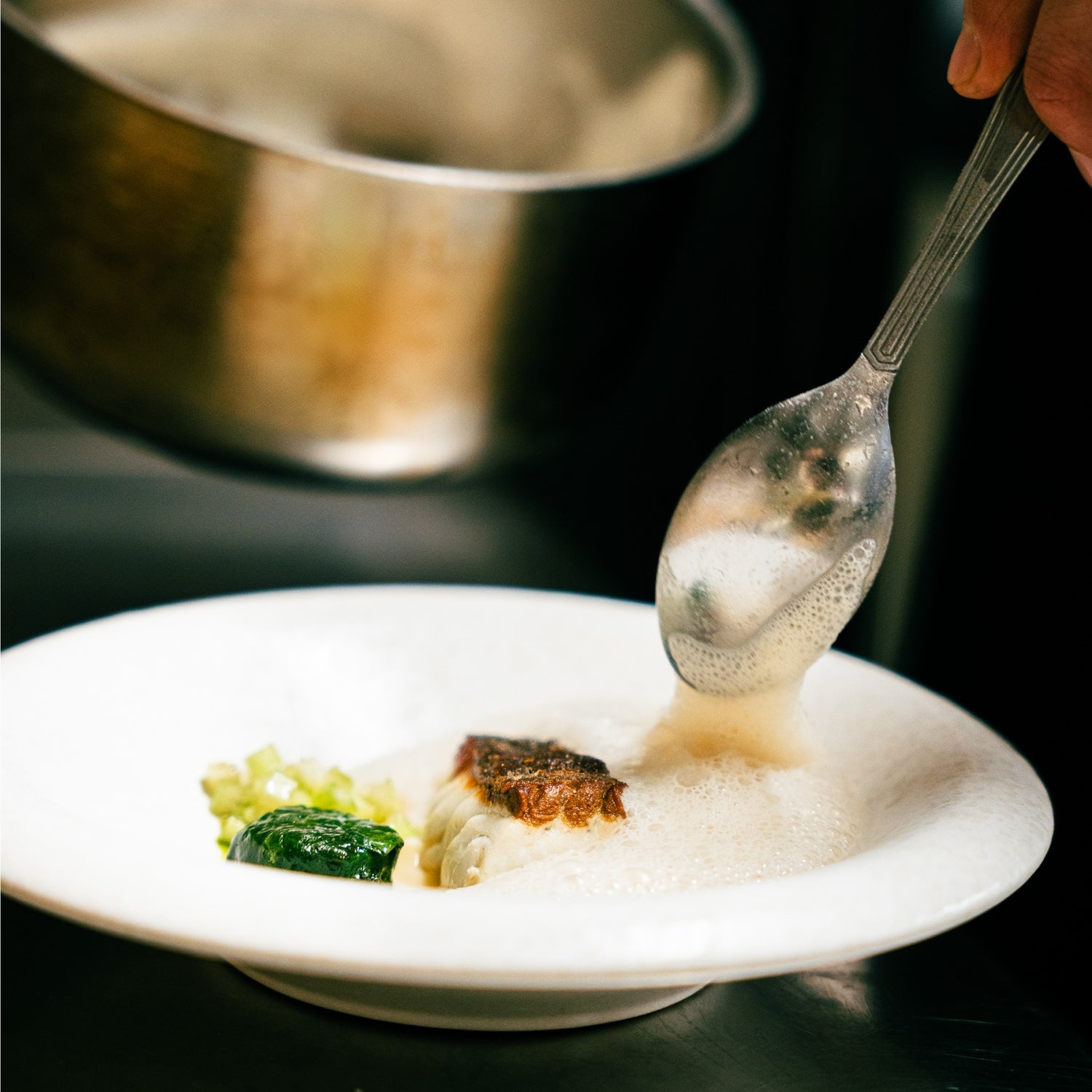 Chef versant délicatement une émulsion mousseuse sur un plat raffiné composé de poisson et de légumes verts.