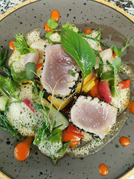 Plat de poisson cru et de légumes, présenté avec une sauce verte et des touches de rouge, dans une assiette grise.