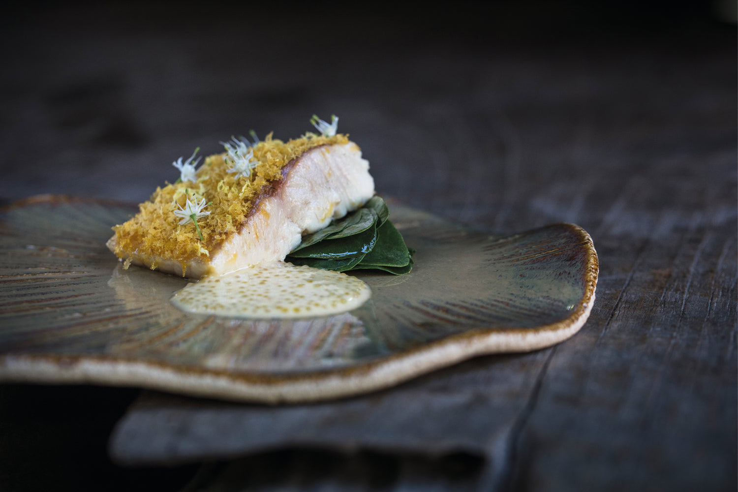 Filet d'omble délicatement pané, garni de fleurs comestibles, posé sur un lit de feuilles vertes et accompagné d'une sauce crémeuse, présenté sur une assiette en céramique texturée.