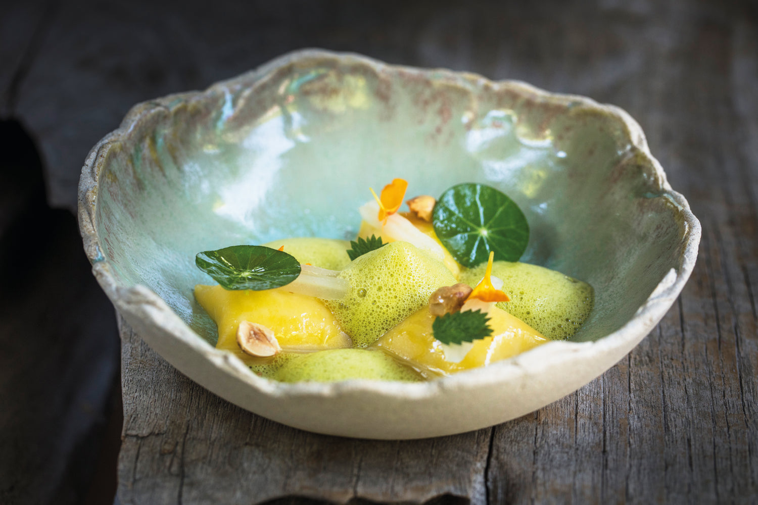 Plat gastronomique de ravioles délicates servies dans une assiette en céramique artisanale, accompagnées d'une mousse verte légère, décorées de feuilles de capucine, de fleurs comestibles et de noisettes croquantes.