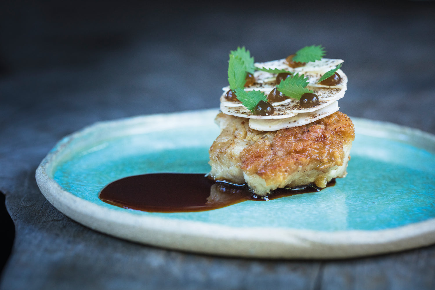 Plat gastronomique mettant en valeur un ris de veau doré, accompagné d'une sauce riche, décoré de fines tranches croustillantes, de touches de sauce et de petites feuilles vertes, servi sur une assiette en céramique turquoise.