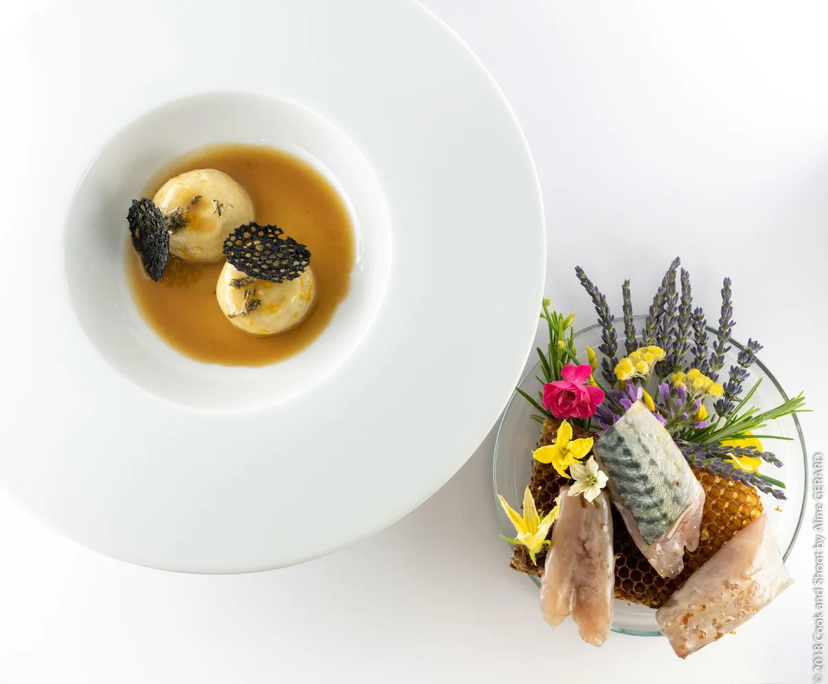 Une assiette blanche contenant un plat sophistiqué : deux petites boules dans une sauce brune, accompagnées de morceaux de poisson et de fleurs disposées artistiquement à côté.