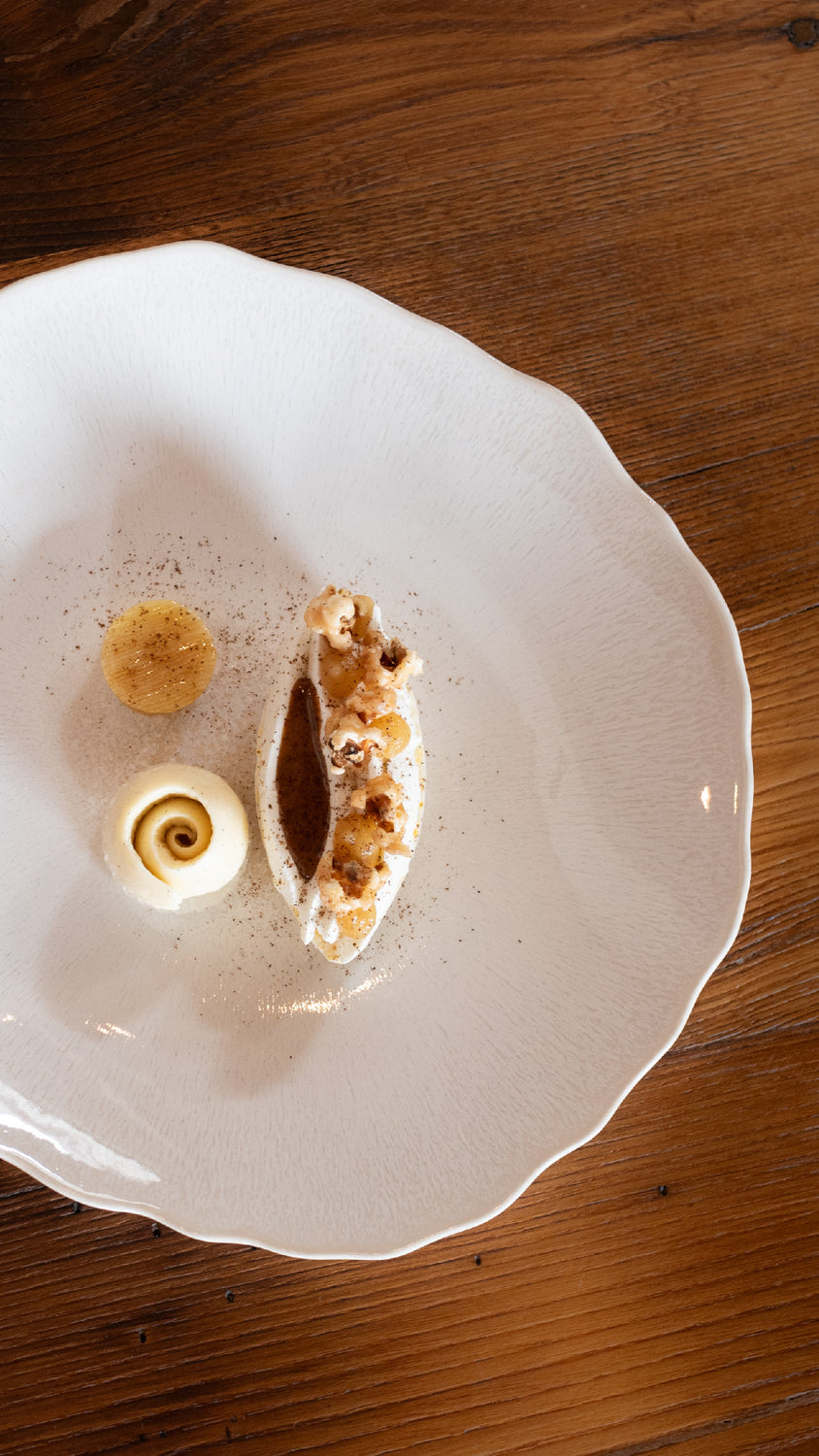 Dessert raffiné présenté dans une assiette blanche texturée, composé d'une mousse légère, d'une spirale de crème vanillée et d'une garniture croquante, disposé avec élégance sur une table en bois.