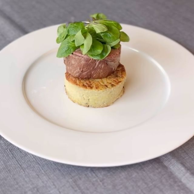 Photo d'un plat de viande surmonté de verdure, présenté sur une base de galette de pommes de terre, le tout disposé sur une assiette blanche.