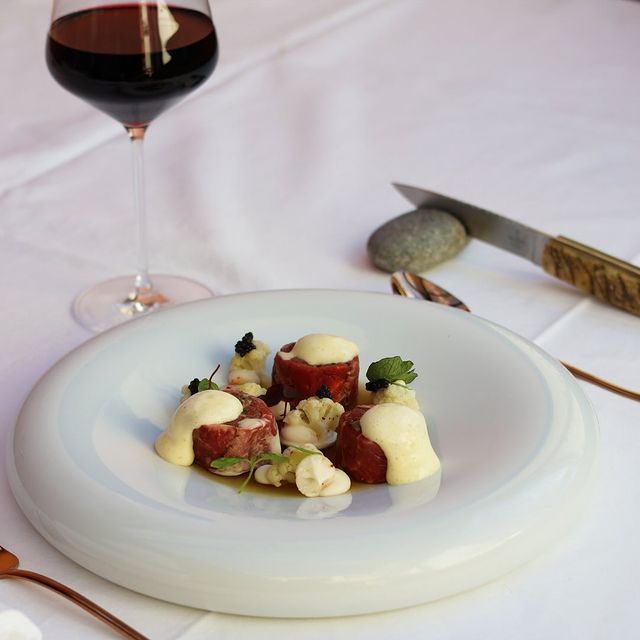 Assiette gastronomique composée de médaillons de viande rouge, garnis de crème et de petites herbes, avec des touches de chou-fleur et de caviar. Un verre de vin rouge accompagne le plat sur une table dressée avec élégance.
