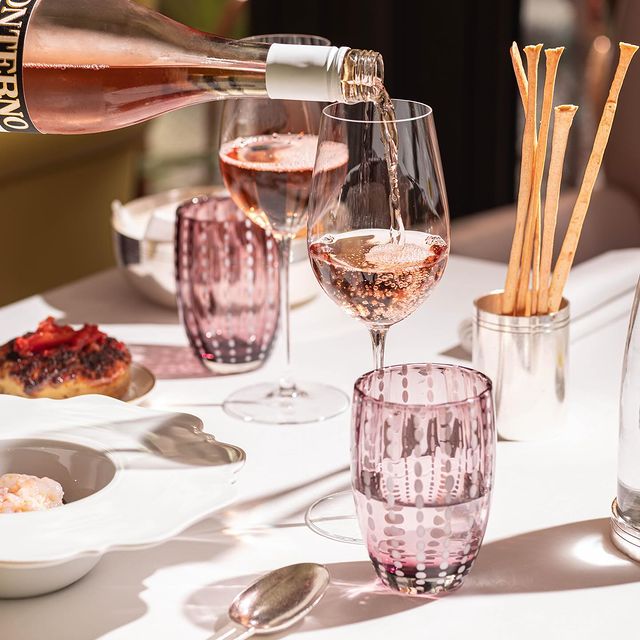 Vin rosé servi dans des verres à vin élégants sur une table dressée pour un repas gastronomique. La table est décorée de verres à motifs roses, de gressins dans un récipient en métal et de plats raffinés. L'ambiance est lumineuse et sophistiquée, parfaite pour une dégustation de vin et un repas élégant.