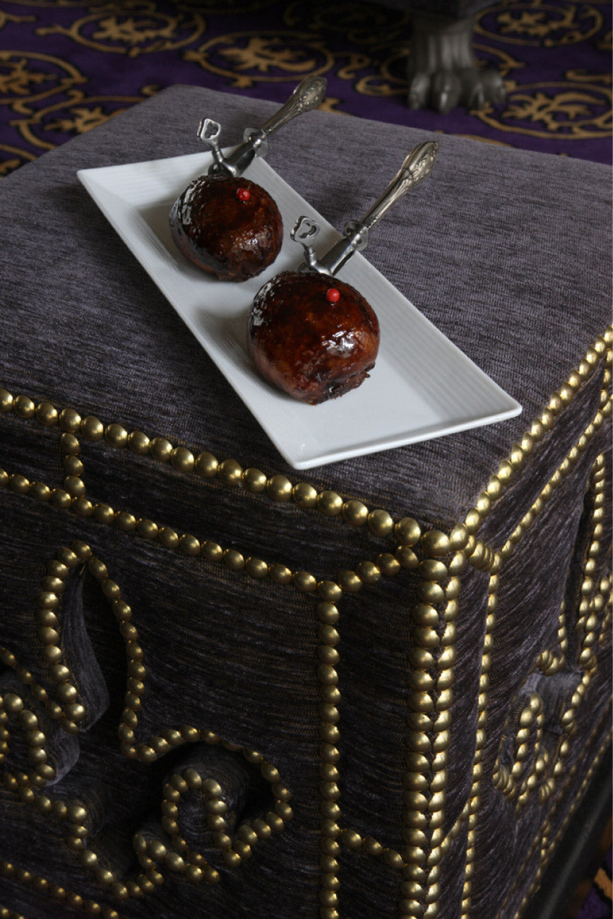 Deux petites pâtisseries glacées au chocolat, chacune surmontée d'une petite bille rouge, présentées sur un plateau rectangulaire blanc avec des couverts argentés, le tout posé sur un meuble en tissu gris orné de clous décoratifs dorés.