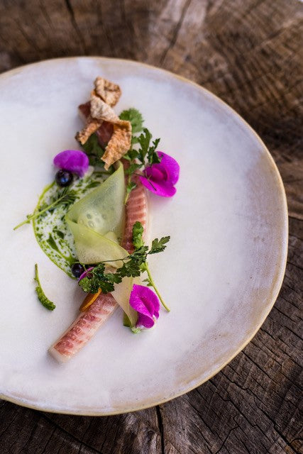 Plat gastronomique délicatement présenté sur une assiette blanche, avec des légumes finement tranchés, des herbes fraîches, des fleurs comestibles violettes, et des morceaux croustillants, le tout disposé de manière artistique.