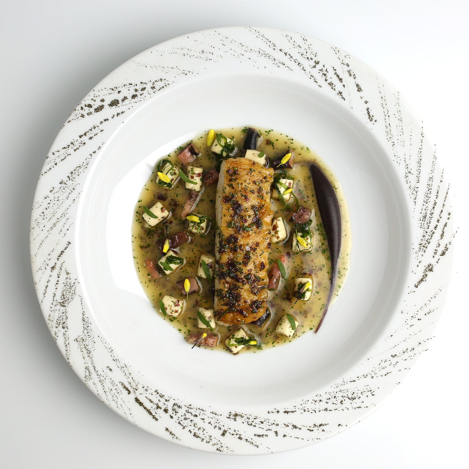 Un plat gastronomique composé d'un filet de poisson parfaitement cuit, servi sur un lit de légumes et accompagné d'une sauce raffinée, le tout présenté dans une assiette blanche ornée de motifs élégants.