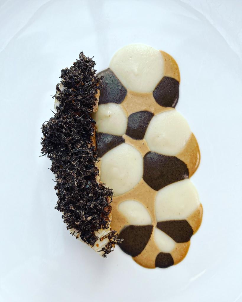 Un plat gastronomique minimaliste avec une pièce de viande ou de poisson recouverte de truffe râpée, accompagnée d'une sauce crémeuse disposée en motifs circulaires bicolores sur une assiette blanche.