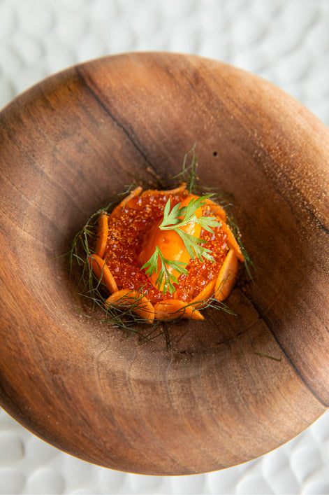 Assiette en bois présentant un plat raffiné avec des œufs de poisson rouges et des herbes fraîches, disposé de manière artistique.