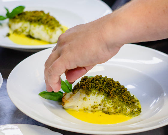 Un plat de poisson garni d'une croûte verte, probablement aux herbes, avec une sauce jaune vif. La main du chef est en train de finaliser la présentation en ajoutant des feuilles vertes fraîches.