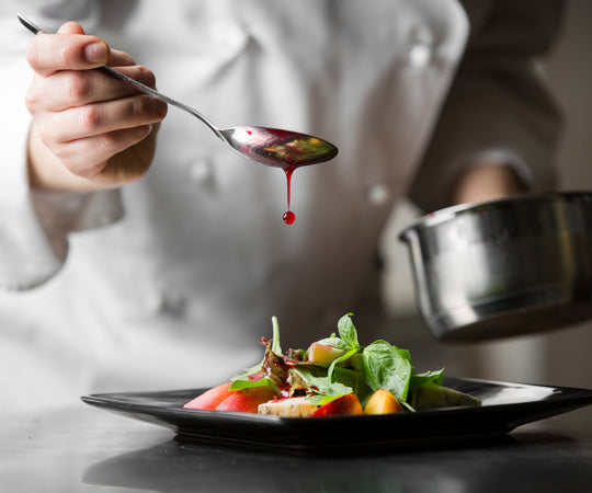 Un chef de cuisine en train de verser délicatement une sauce rouge sur une salade, montrant une attention minutieuse aux détails culinaires.