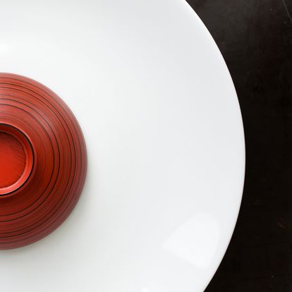 Un bol rouge laqué posé à l'envers sur une grande assiette blanche minimaliste, créant un contraste net sur un fond sombre.