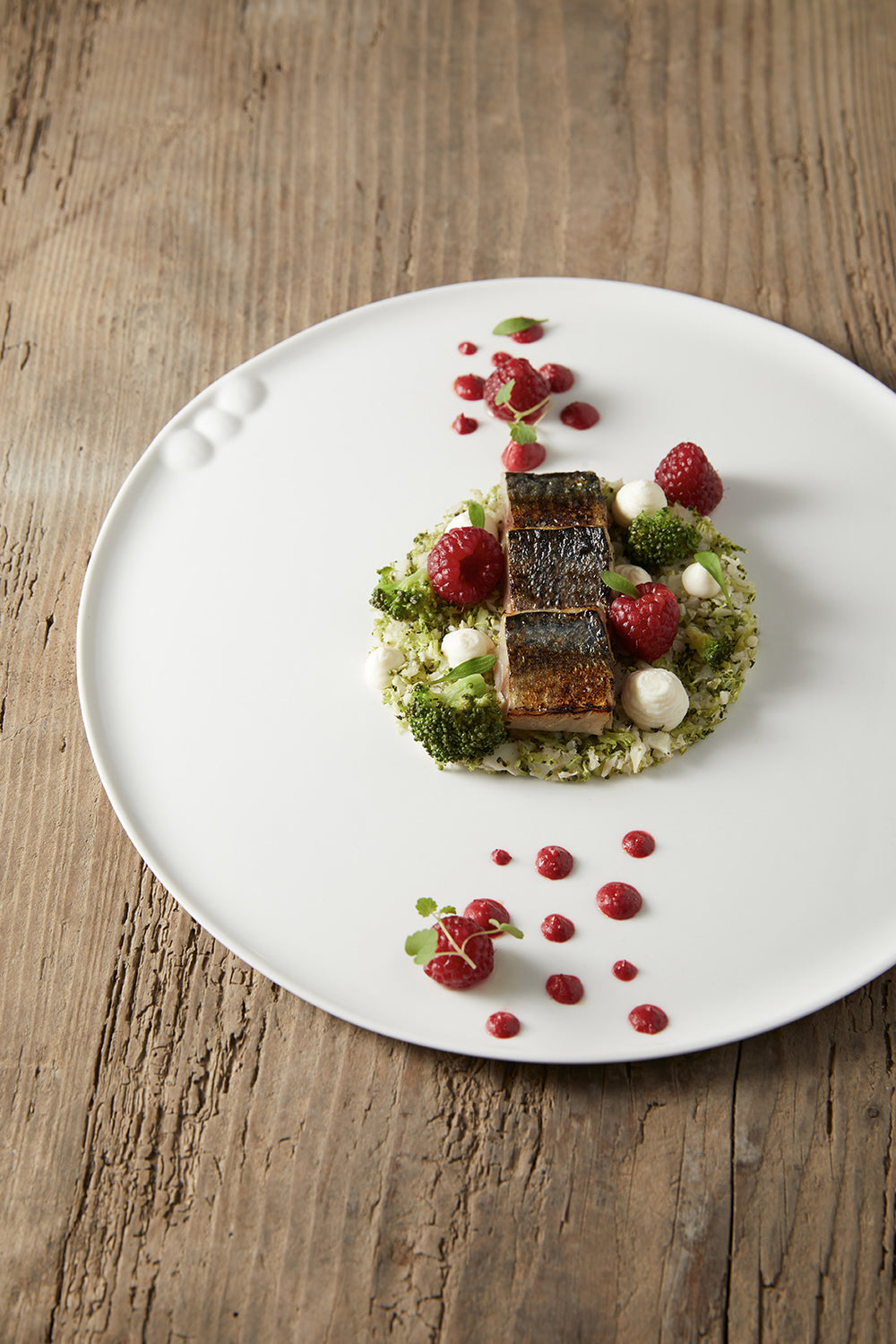 Plat gastronomique composé de poisson grillé, de framboises et de légumes, joliment disposé sur une assiette blanche.
