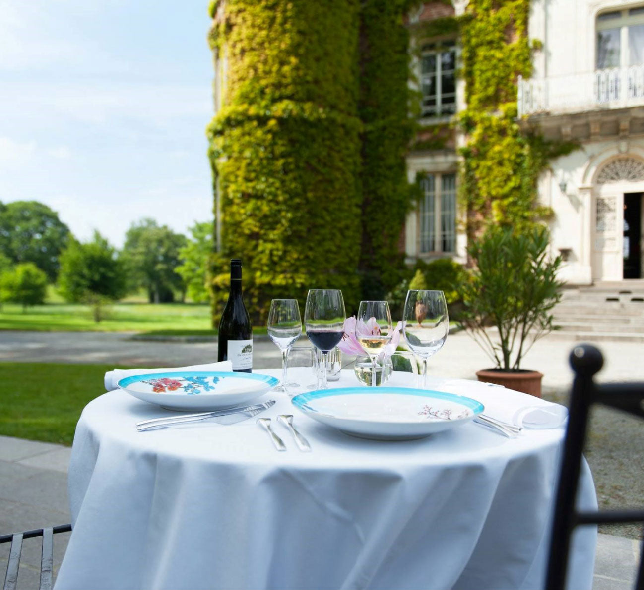 Table dressée en extérieur devant un manoir recouvert de lierre, avec verres à vin et assiettes décorées.