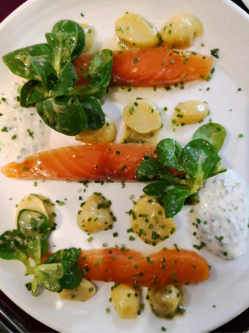 Assiette de saumon mariné avec de petites pommes de terre, des feuilles de mâche, et une sauce crémeuse à l'herbe, le tout décoré de ciboulette hachée.