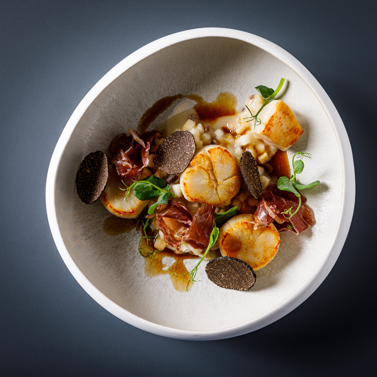 Plat gastronomique élégant avec des noix de Saint-Jacques dorées, accompagnées de truffes noires, de jambon cru et d'une garniture crémeuse dans un bol raffiné.