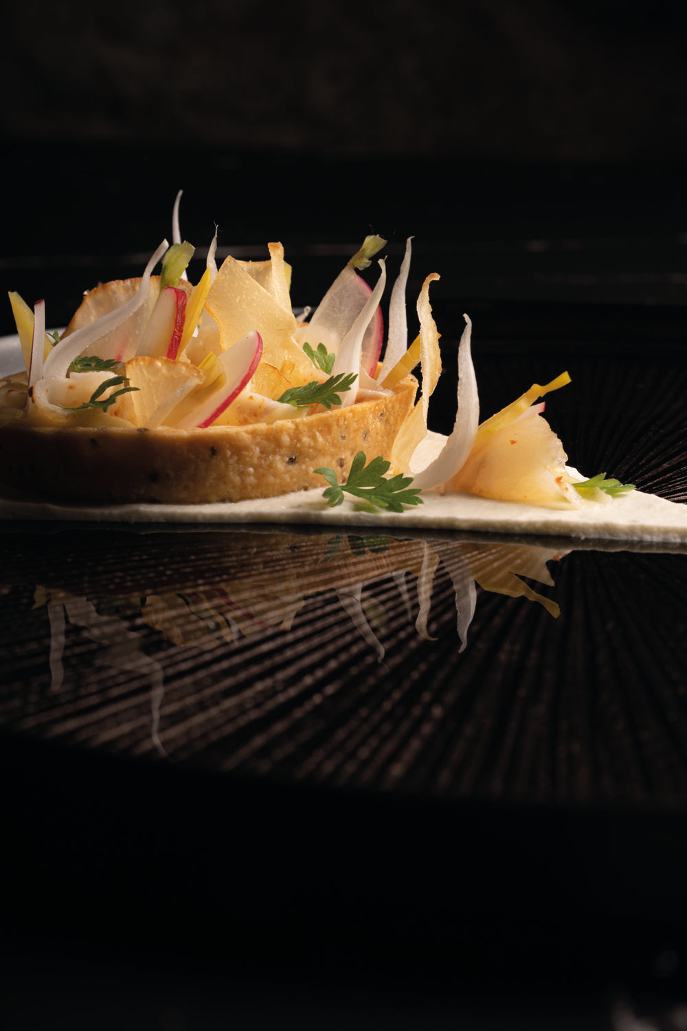 Plat gastronomique délicatement présenté avec des légumes finement tranchés disposés en éventail sur une base croustillante, le tout accompagné d'herbes fraîches et éclairé d'une lumière douce, créant une ambiance élégante et raffinée.