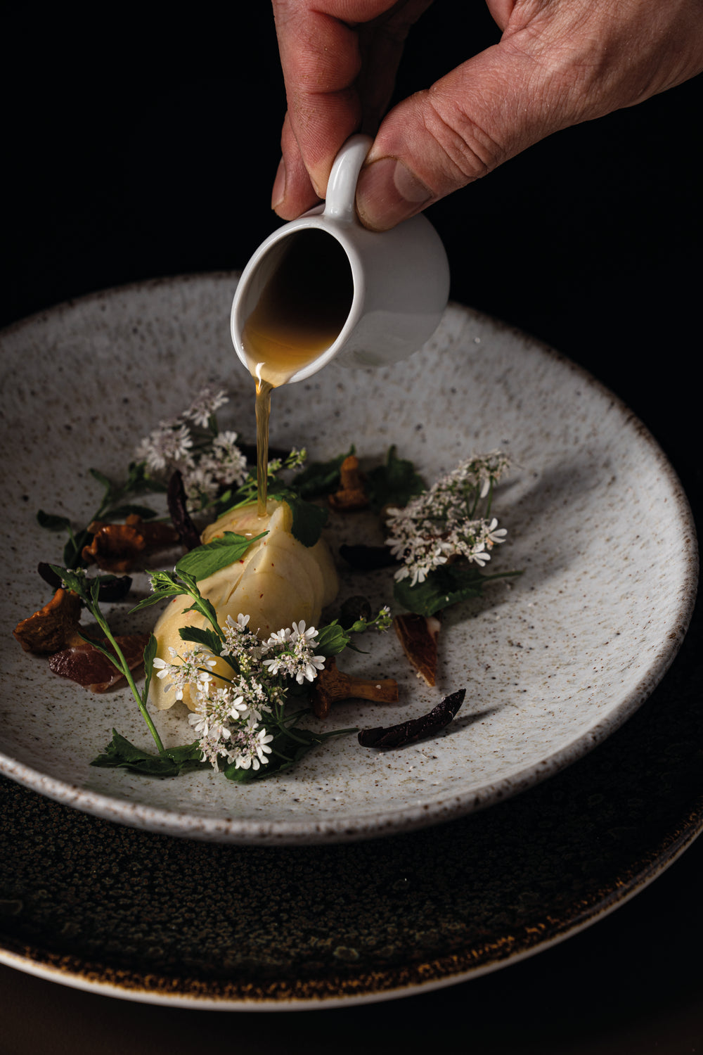 Un plat raffiné dans une assiette en céramique, garni de fleurs comestibles et de verdure, avec une quenelle au centre. Un filet de sauce est délicatement versé sur le plat à partir d'un petit pot blanc, mettant en valeur l'élégance et la subtilité de la présentation.