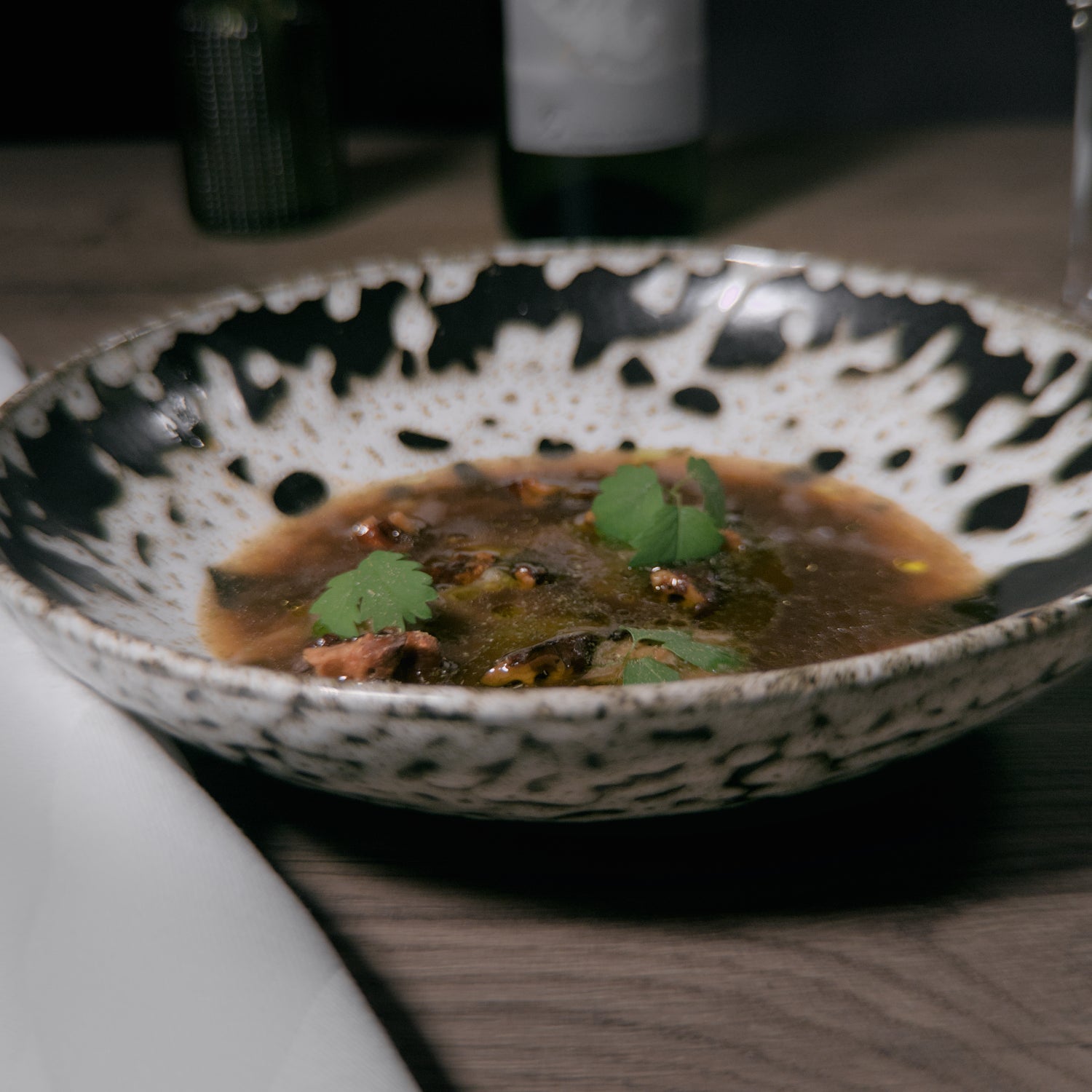 Plat gastronomique présenté dans une assiette en céramique blanche et noire, contenant une soupe ou un ragoût avec des morceaux de viande et des herbes fraîches. La table est dressée avec une serviette blanche et un verre à vin en arrière-plan. L'ambiance est élégante et raffinée.