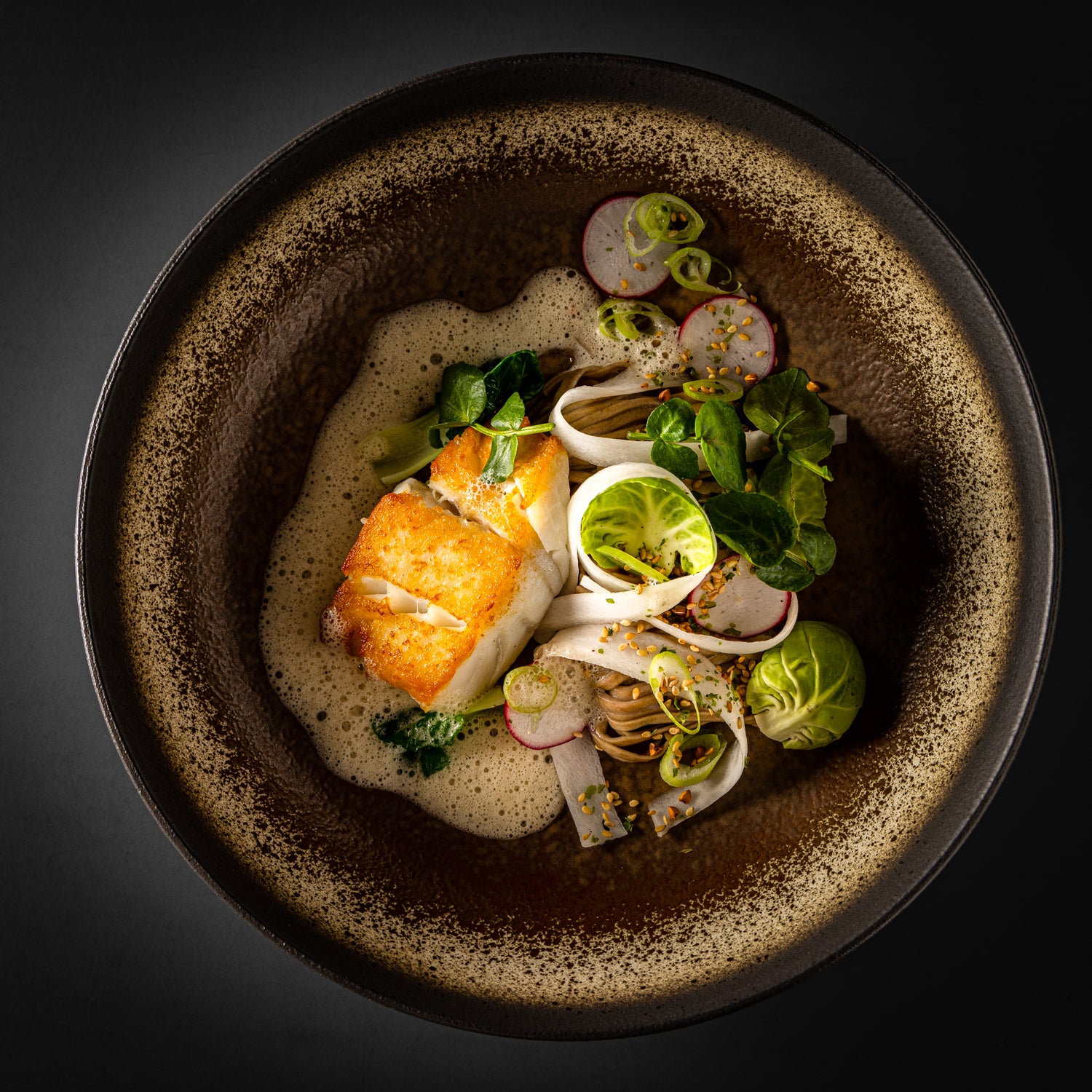 Plat gastronomique dans un bol élégant, composé de poisson poêlé, de légumes frais tels que radis et choux de Bruxelles, sur un lit de nouilles soba avec une mousse légère.