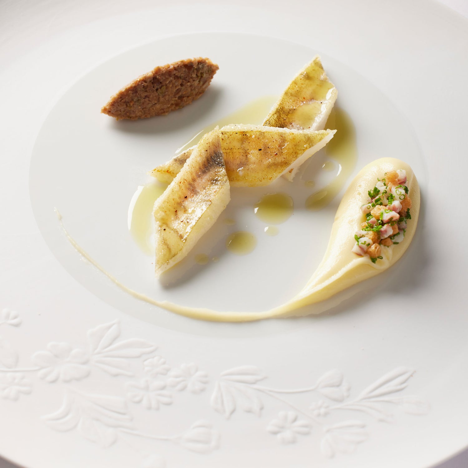 Assiette gastronomique élégante avec un plat de poisson délicatement dressé, accompagné d'une quenelle de purée et d'une garniture raffinée, sur une assiette blanche ornée de motifs floraux.