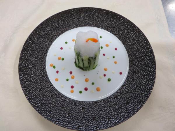 Assiette gastronomique élégante avec un plat vertical composé de légumes verts, surmonté d'une mousse blanche, entouré de petites gouttes de sauces colorées sur un fond blanc, le tout disposé sur une grande assiette noire texturée.