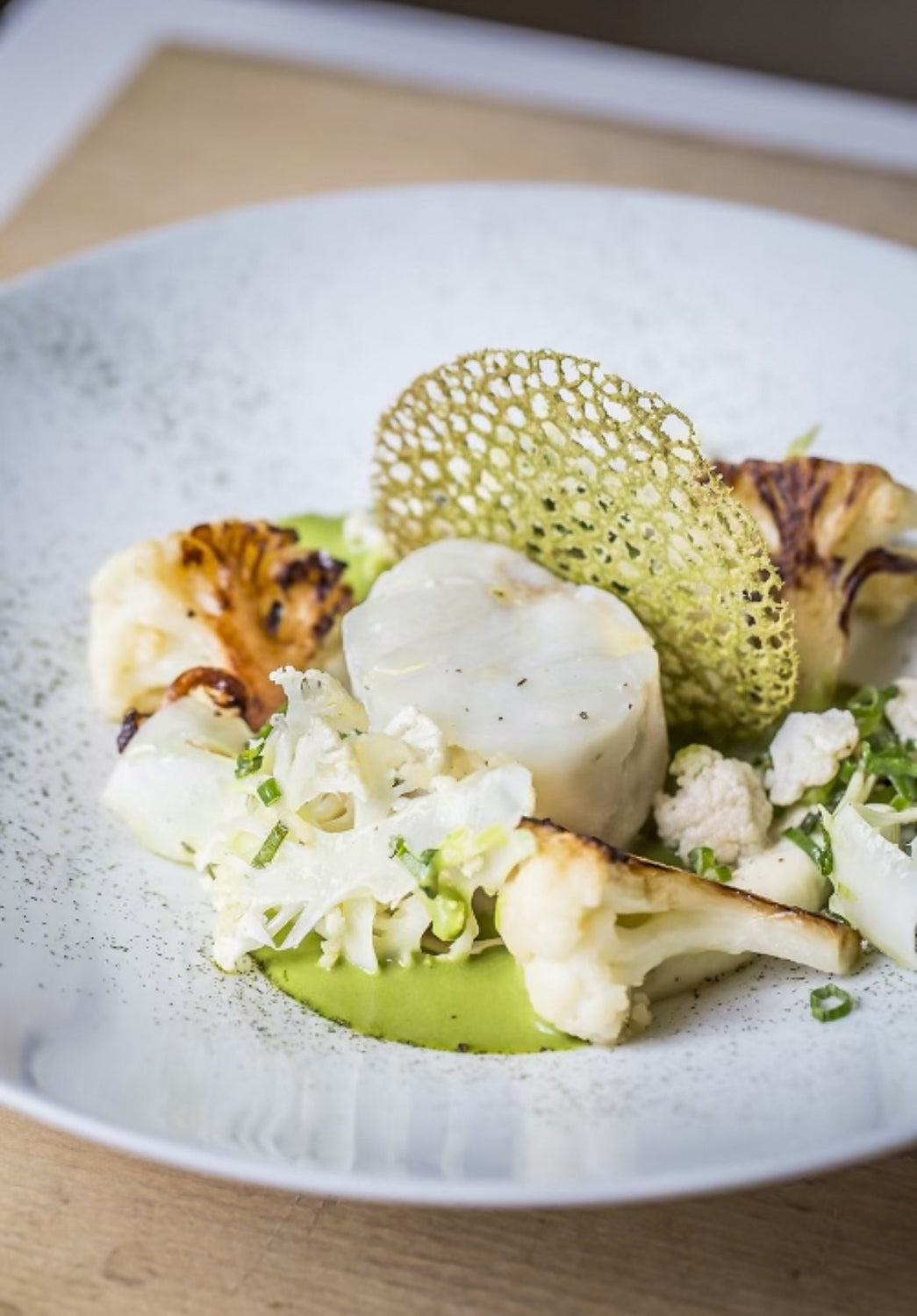 Assiette gastronomique avec un pavé de poisson blanc, accompagné de choux-fleurs grillés et crus, avec une sauce verte et une tuile croustillante verte.