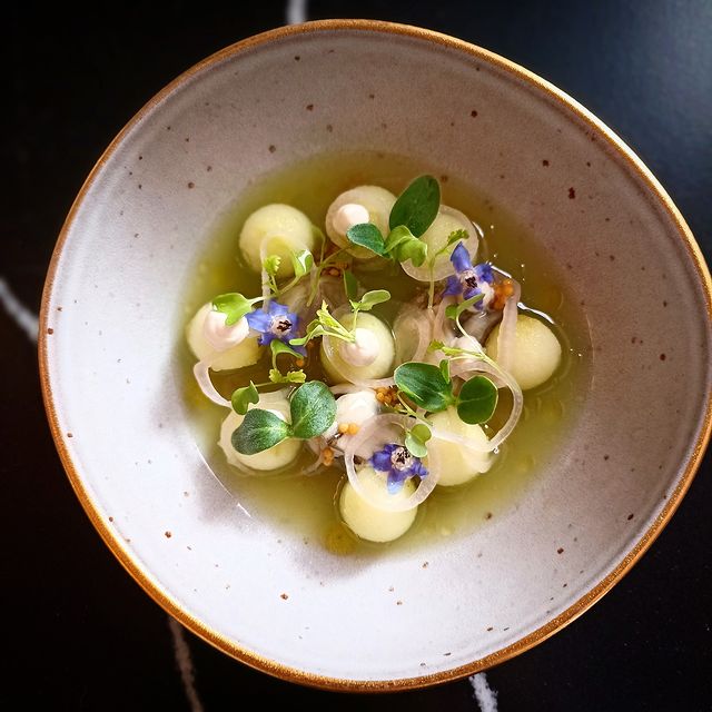 Bouillon aux billes de melon, herbes fraîches et fleurs comestibles dans une assiette profonde au design élégant.
