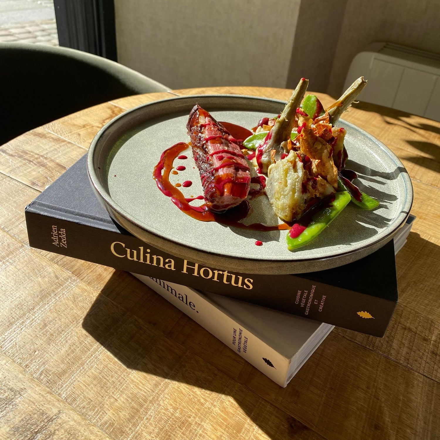 Plat gastronomique dressé sur une assiette, posée sur deux livres empilés intitulés 'Culina Hortus' et 'Animale', avec une sauce rouge éclatante et des légumes artistiquement présentés.