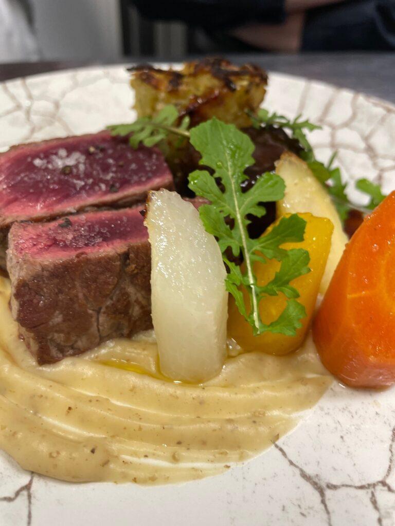 Une assiette montrant un morceau de viande rosée servi sur une purée crémeuse, accompagné de légumes cuits, avec une présentation soignée et colorée.