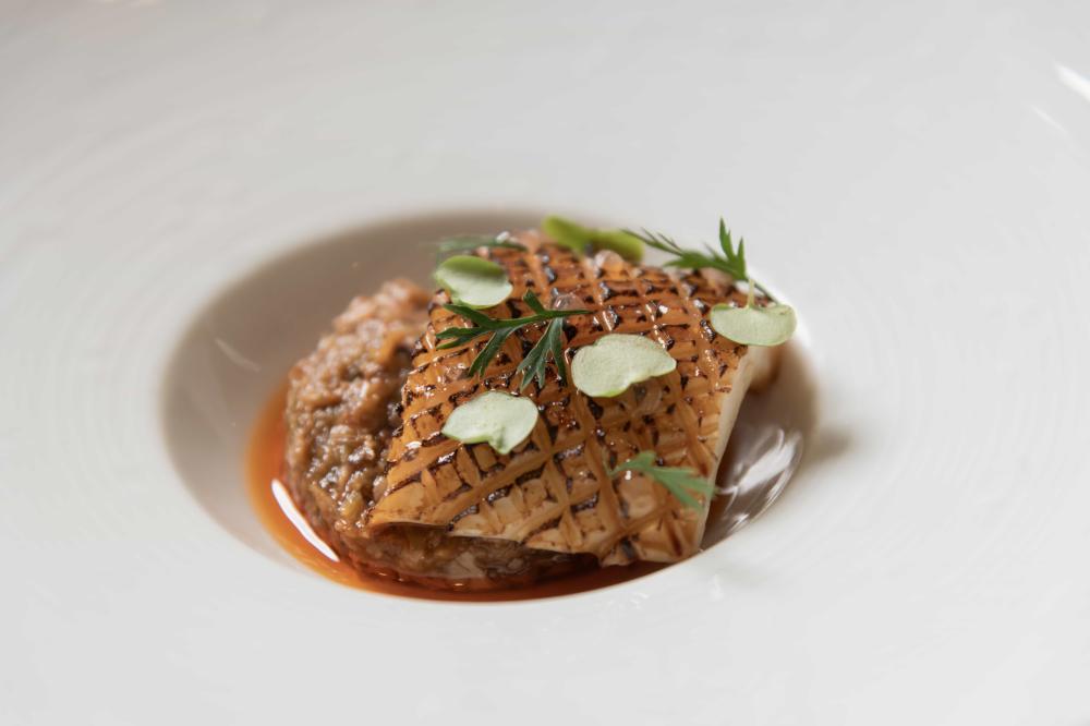 Plat gastronomique avec un morceau de poisson délicatement grillé et marqué de motifs en croisillons, servi sur un lit de risotto crémeux, agrémenté de petites feuilles vertes et d'une sauce riche.