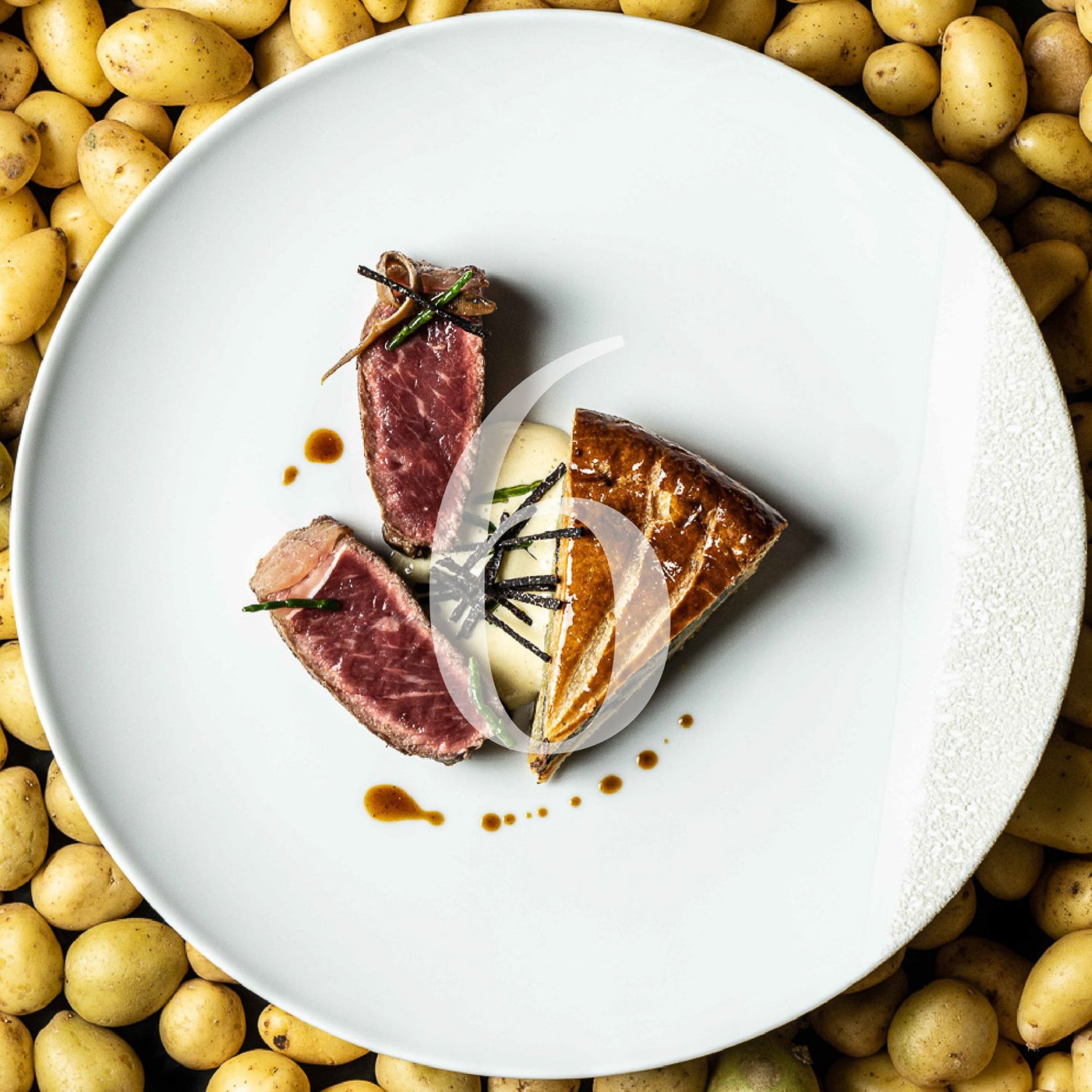 Plat gastronomique raffiné présenté dans une assiette blanche, composé de deux morceaux de viande rouge saignante, d'une garniture crémeuse avec des copeaux noirs et d'une part de tourte dorée, le tout disposé sur un fond de petites pommes de terre jaunes.