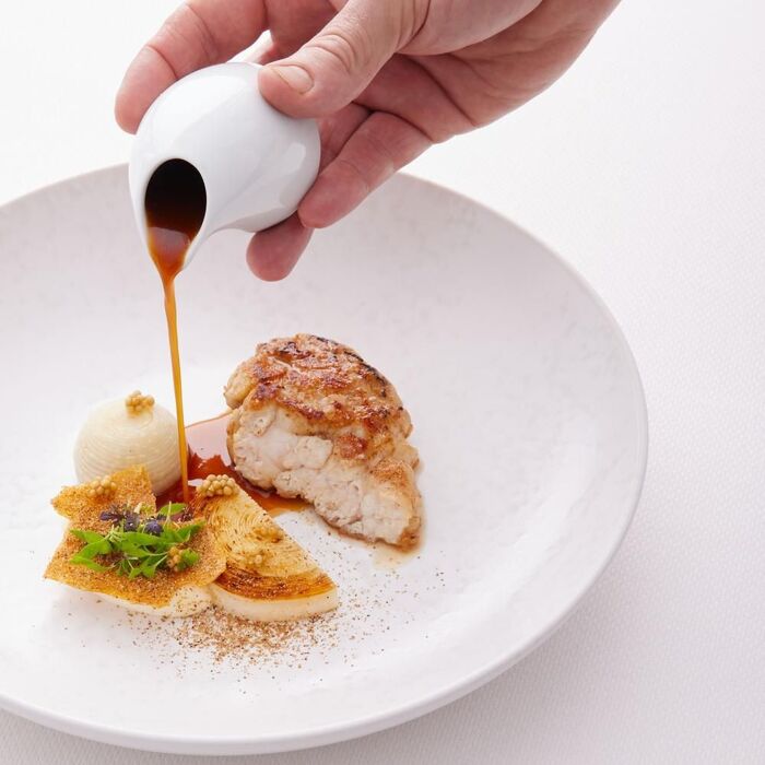 Un plat raffiné où une main verse délicatement une sauce sur une pièce de viande, accompagnée de garnitures artistiquement disposées, dans une assiette blanche texturée.