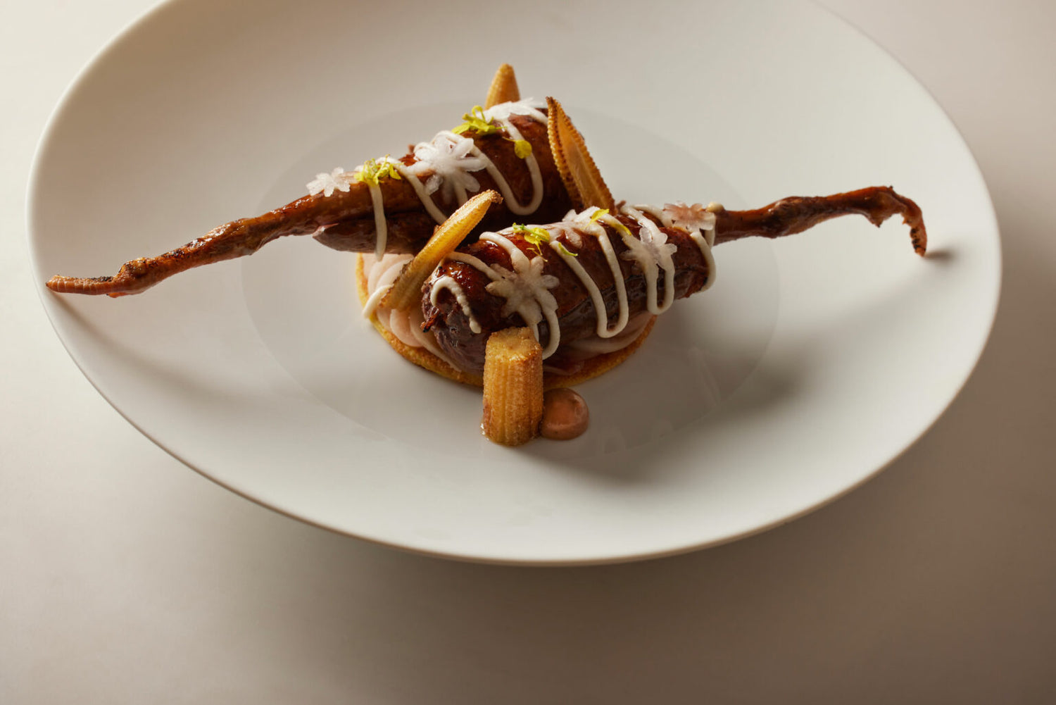 Un plat innovant où une pièce de viande est dressée sur une base circulaire, entourée de maïs miniatures et d'une sauce raffinée, servi dans une grande assiette blanche.