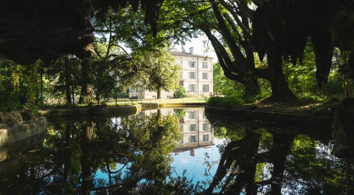 Vue de loin sur le restaurant, grand domaine avec un lac en premier plan sur lequel les arbres se reflètent 