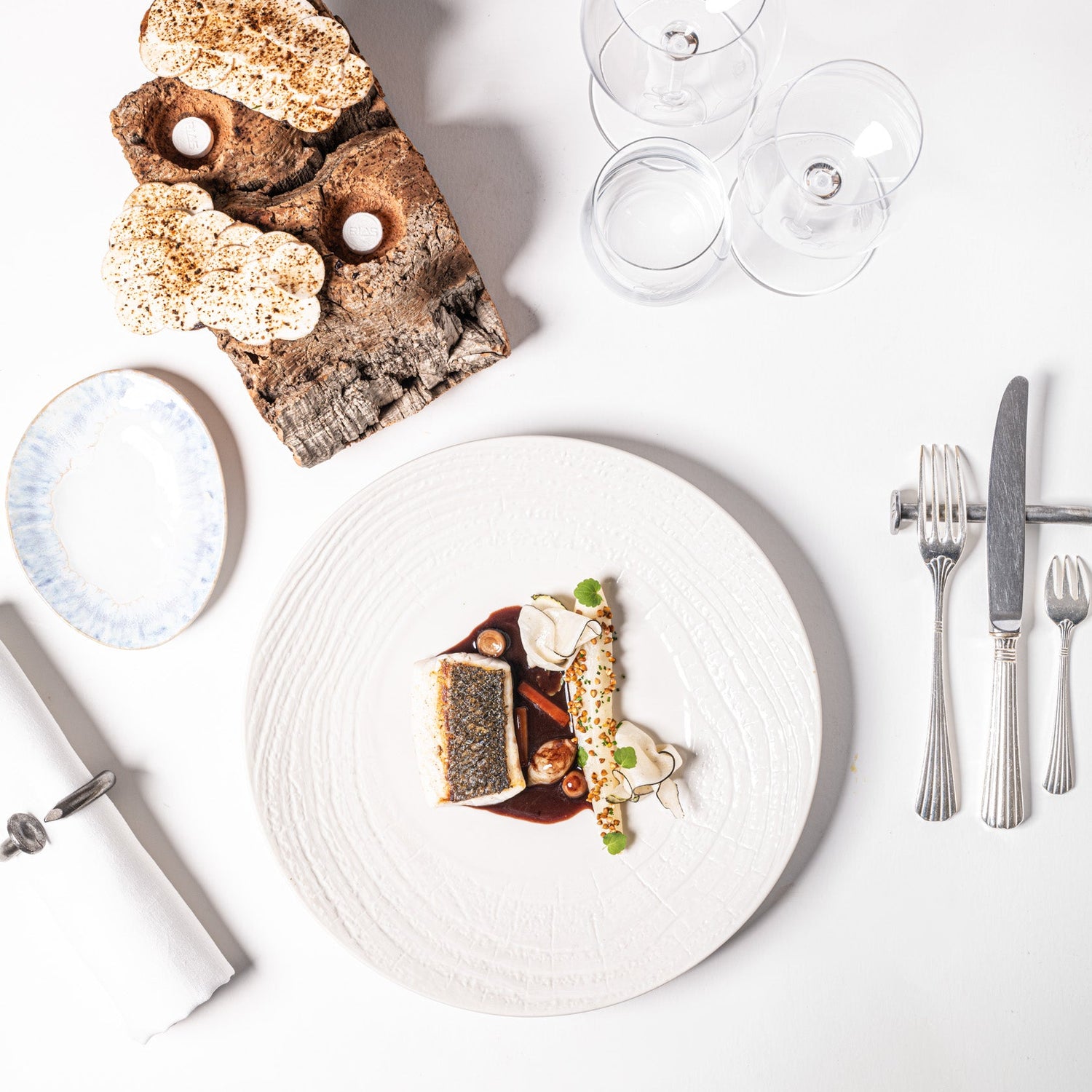 Assiette élégante avec une variété de petits plats gastronomiques, accompagnée de couverts et de verres à vin sur une table blanche.