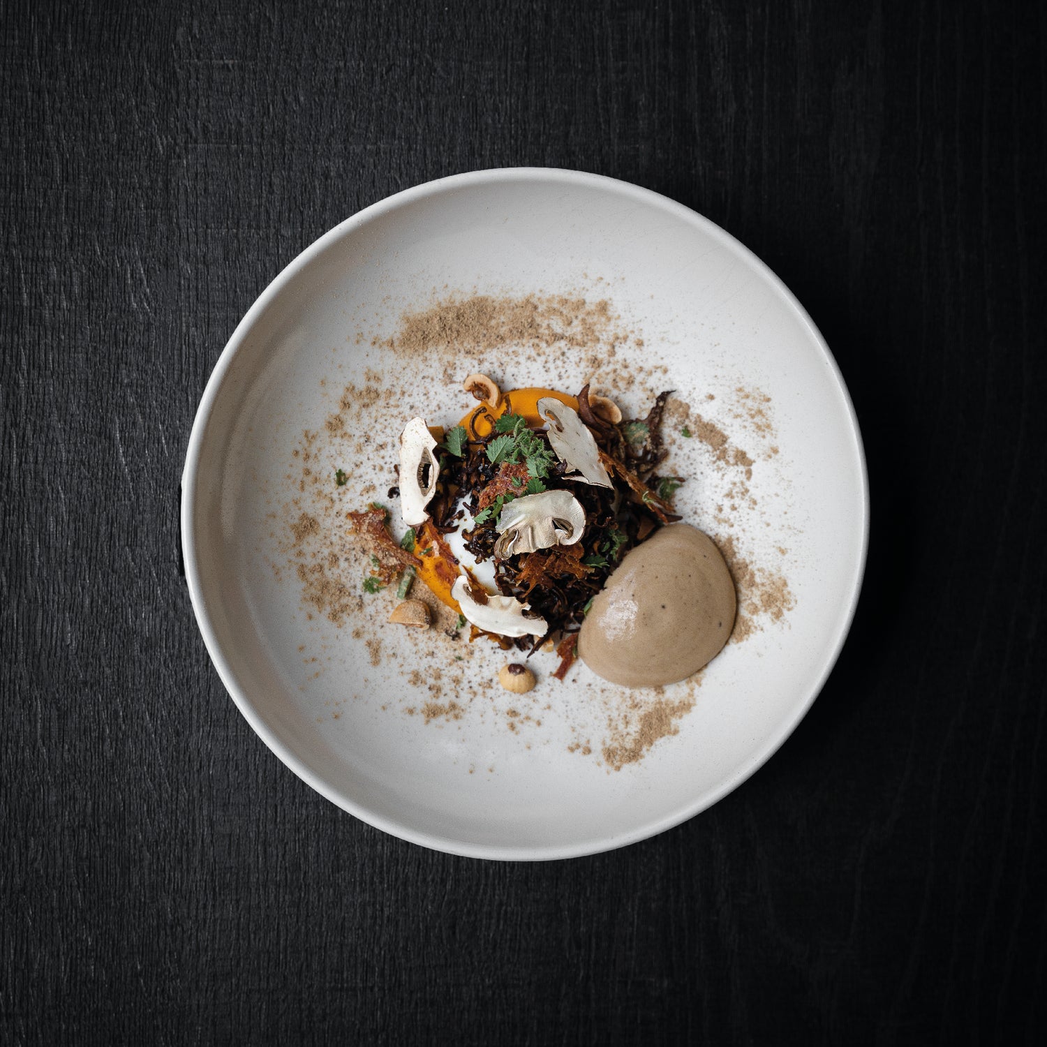 Plat gastronomique élégant présenté dans une assiette blanche, avec des légumes, des champignons et une mousse délicatement disposée, saupoudré d'épices.