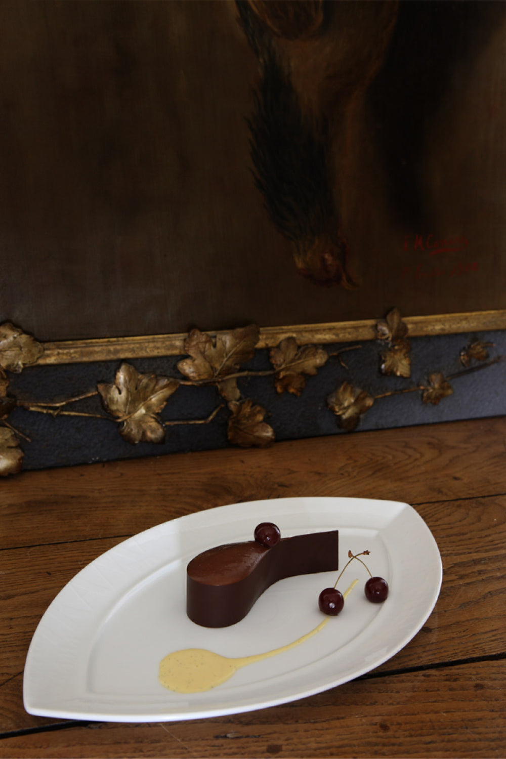 Dessert chocolaté en forme élégante avec des cerises et une sauce à la vanille sur une assiette blanche, présenté devant un tableau artistique.