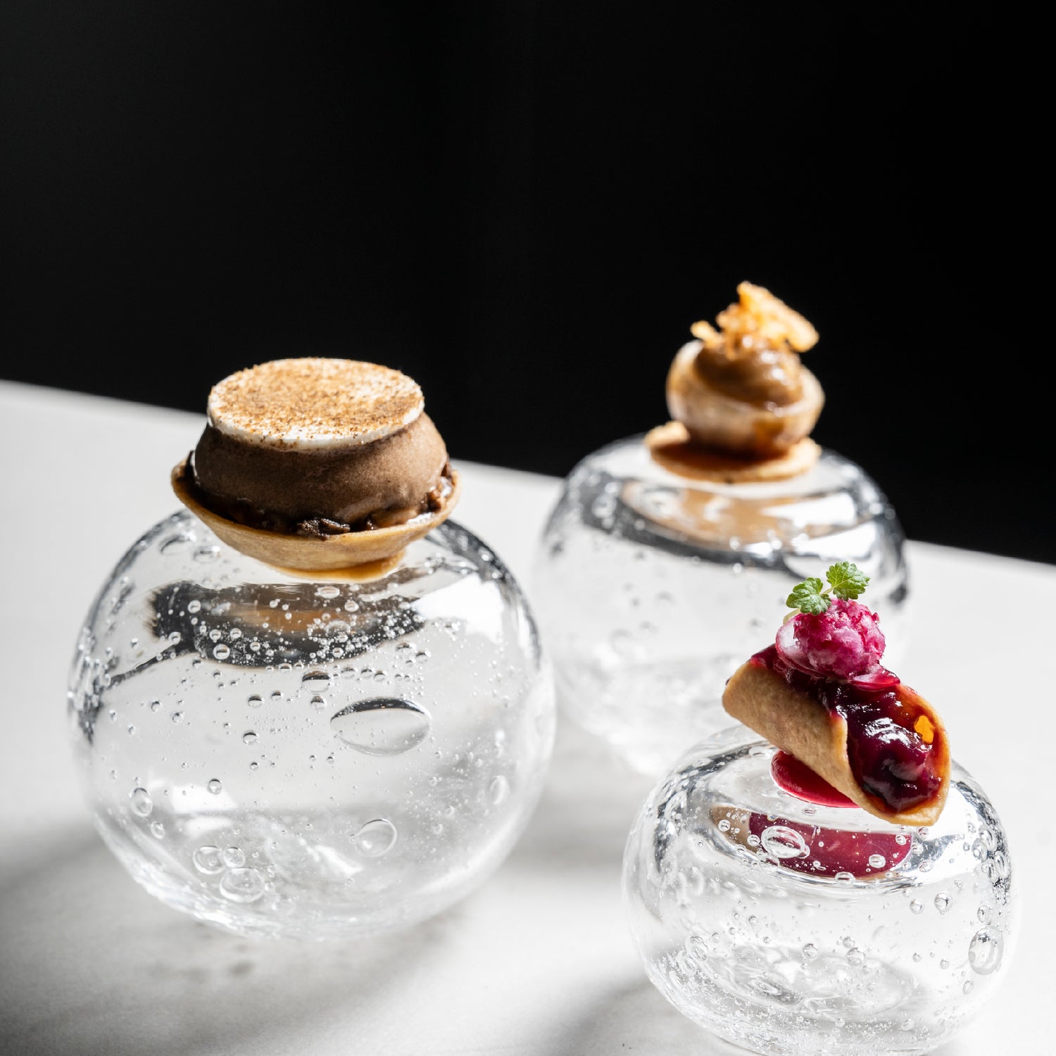 Assortiment de mignardises raffinées présentées sur des supports en verre soufflé transparent.