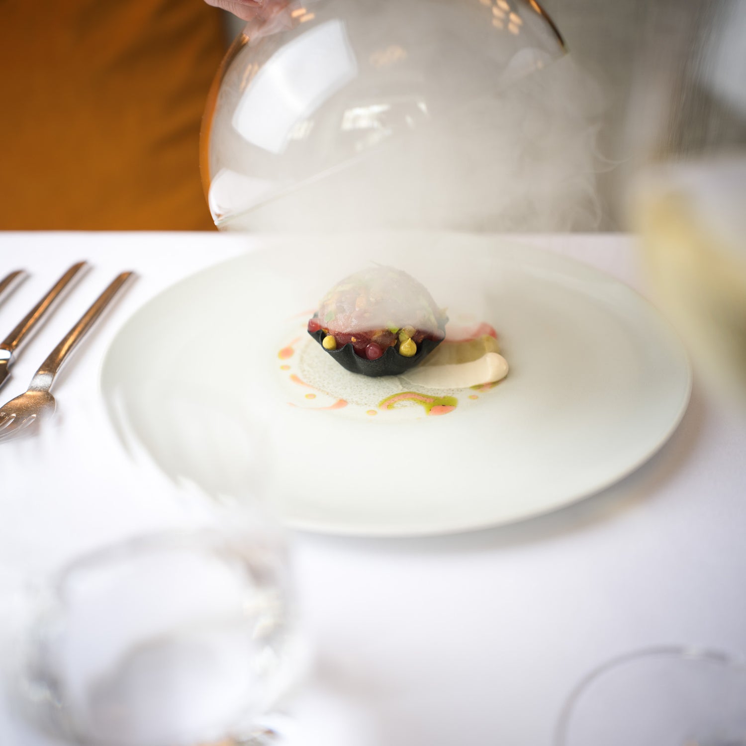 Un plat gastronomique servi sous une cloche en verre, dévoilant une préparation délicate avec des touches de couleur, entouré de vapeur pour un effet spectaculaire.