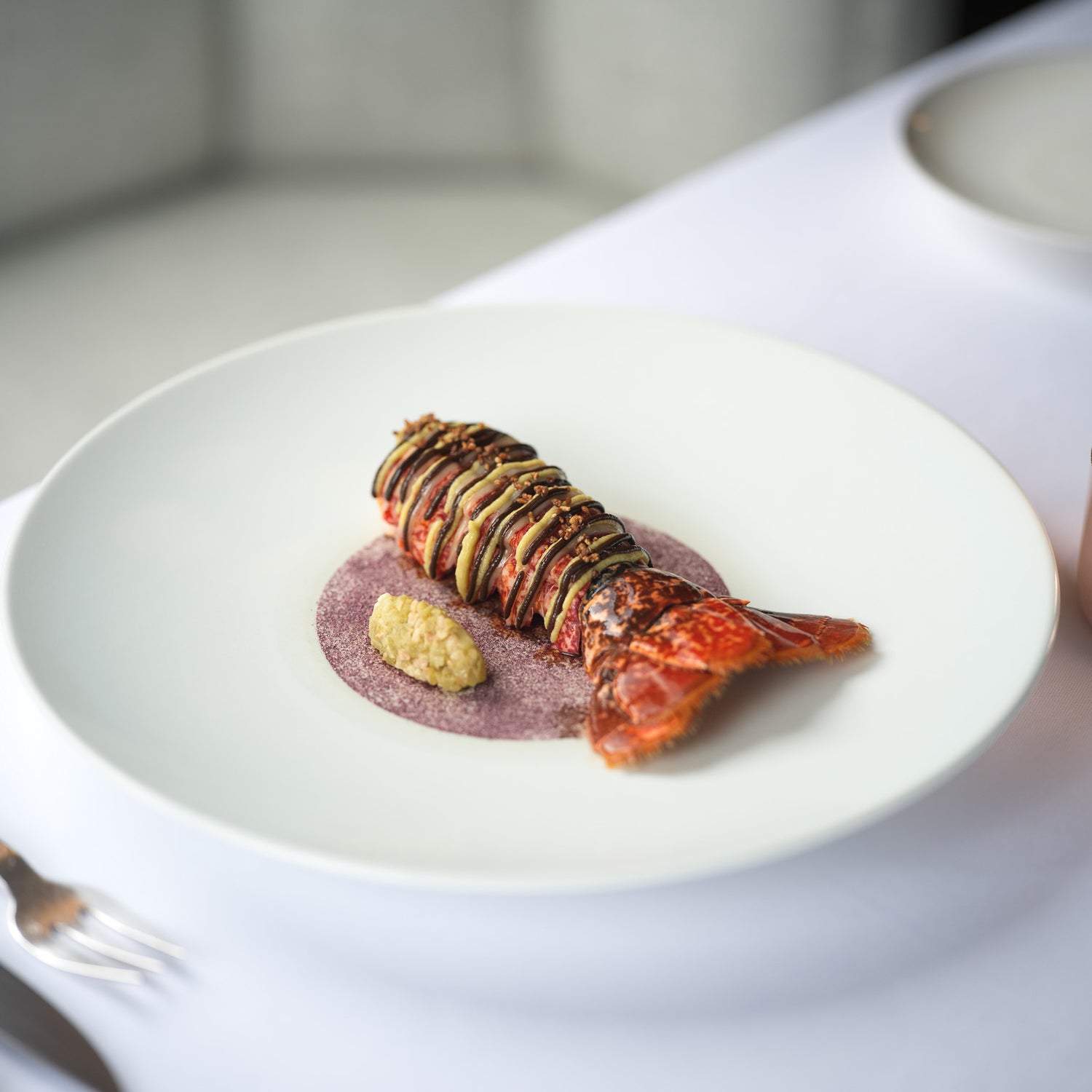 Plat gastronomique composé d'une queue de homard finement dressée avec des légumes en fines lamelles sur une sauce colorée, servi dans une assiette élégante avec une petite casserole en cuivre contenant une sauce à côté.