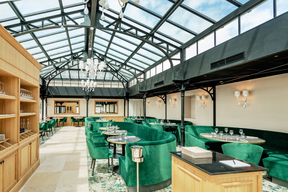 Salle de restaurant élégante sous une verrière lumineuse, avec des banquettes et des fauteuils en velours vert, des tables raffinées et une décoration mêlant bois clair et métal noir dans un style contemporain et sophistiqué.