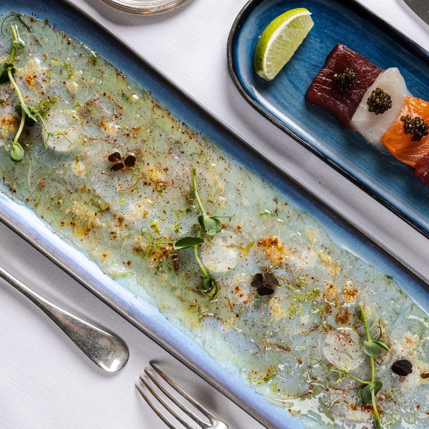 Assiettes de poissons crus finement tranchés, carpaccio à gauche et sashimi avec caviar à droite, présentées dans des plats bleus.